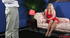 Busty blonde wearing sexy red dress and silver high heels sits on a pink couch while talking to a guy on how she likes a man in boxers.
